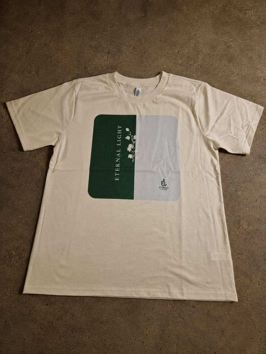 The premium ecru cream graphic t-shirt laid out flat on brown concrete floor.