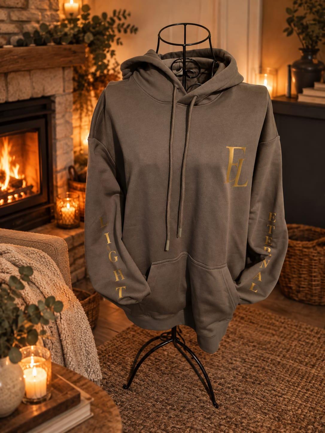 Charcoal green Eternal Light hoodie displayed on a dress form beside a lit fireplace, showing its gold chest monogram and sleeve lettering.