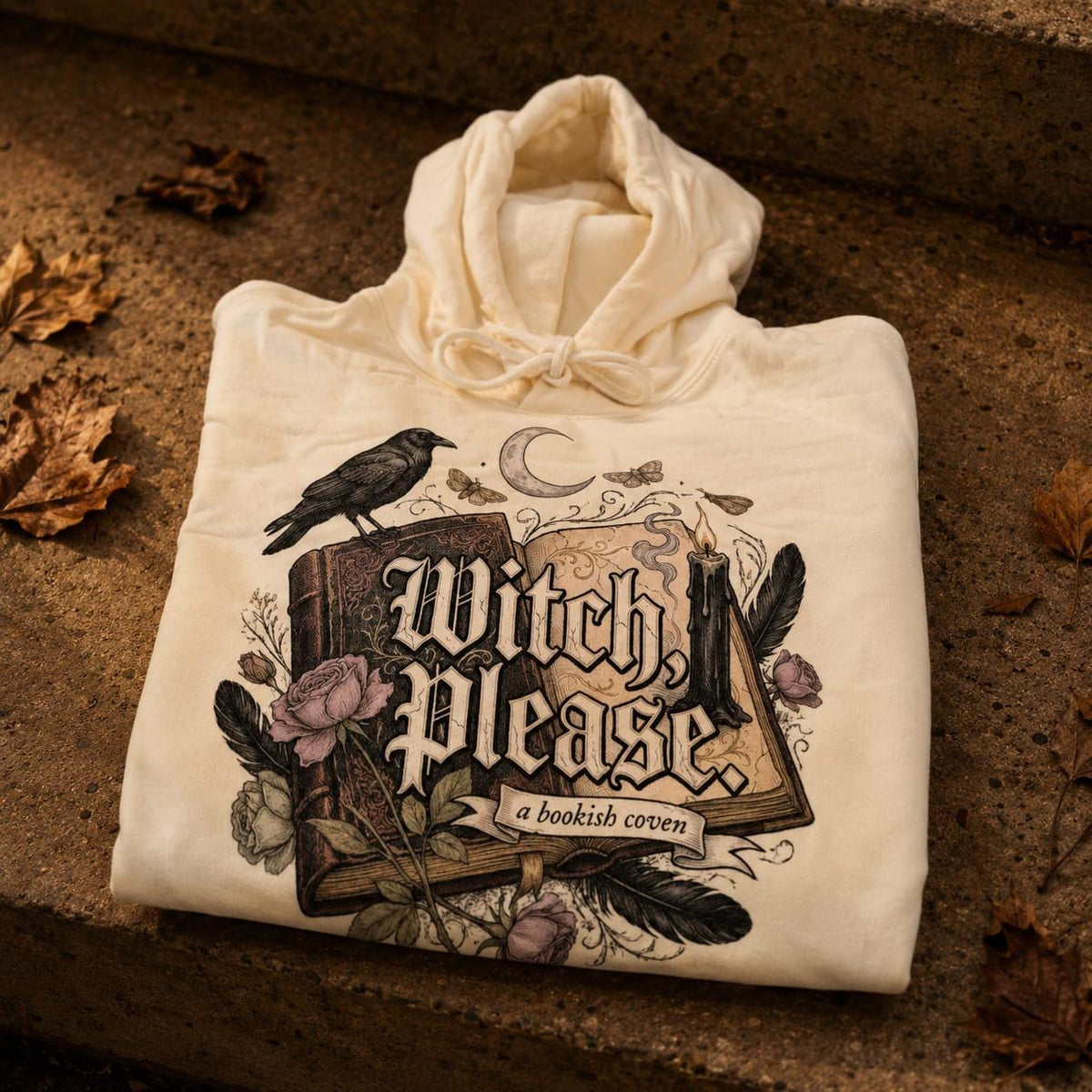 A folded, premium heavyweight hoodie in a soft, natural Rice Apricot cream colour, laid flat on a rustic surface. The front of the garment features an intricate, hand-drawn dark academia illustration. 