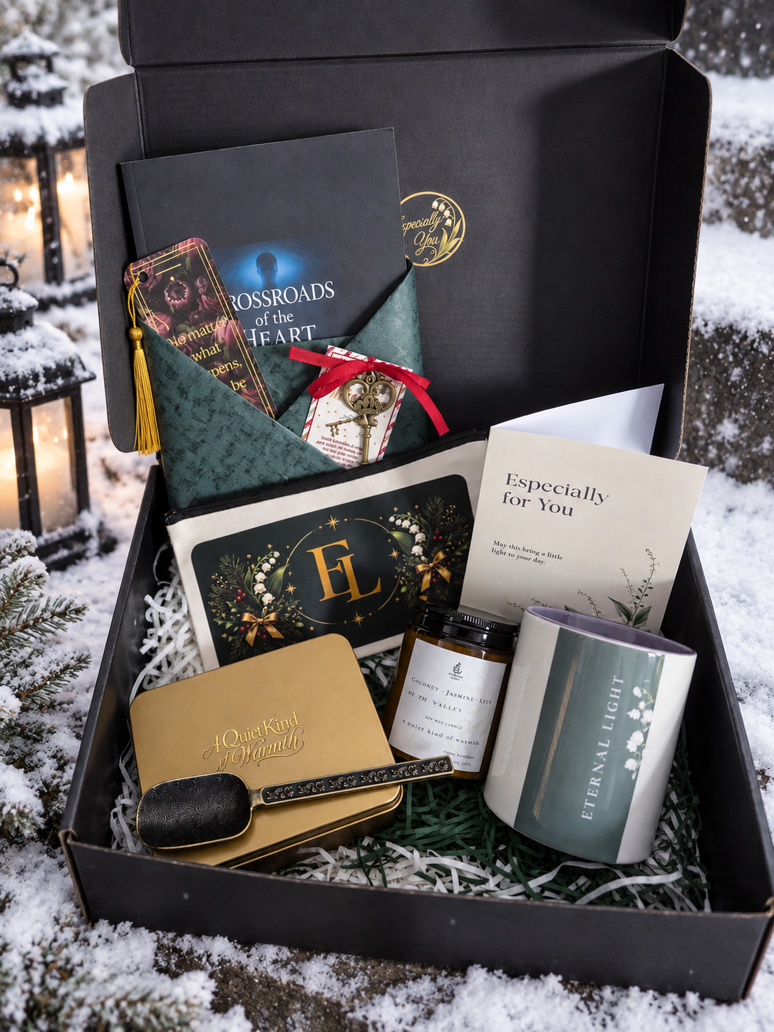 A Christmas-themed gift box filled with cozy items, including a luxury candle, a signed Crossroads of the Heart novel, a pouch, a mug, and a small decorative gold tin filled with goodies, arranged in shredded paper inside a black box surrounded by snowy elements.