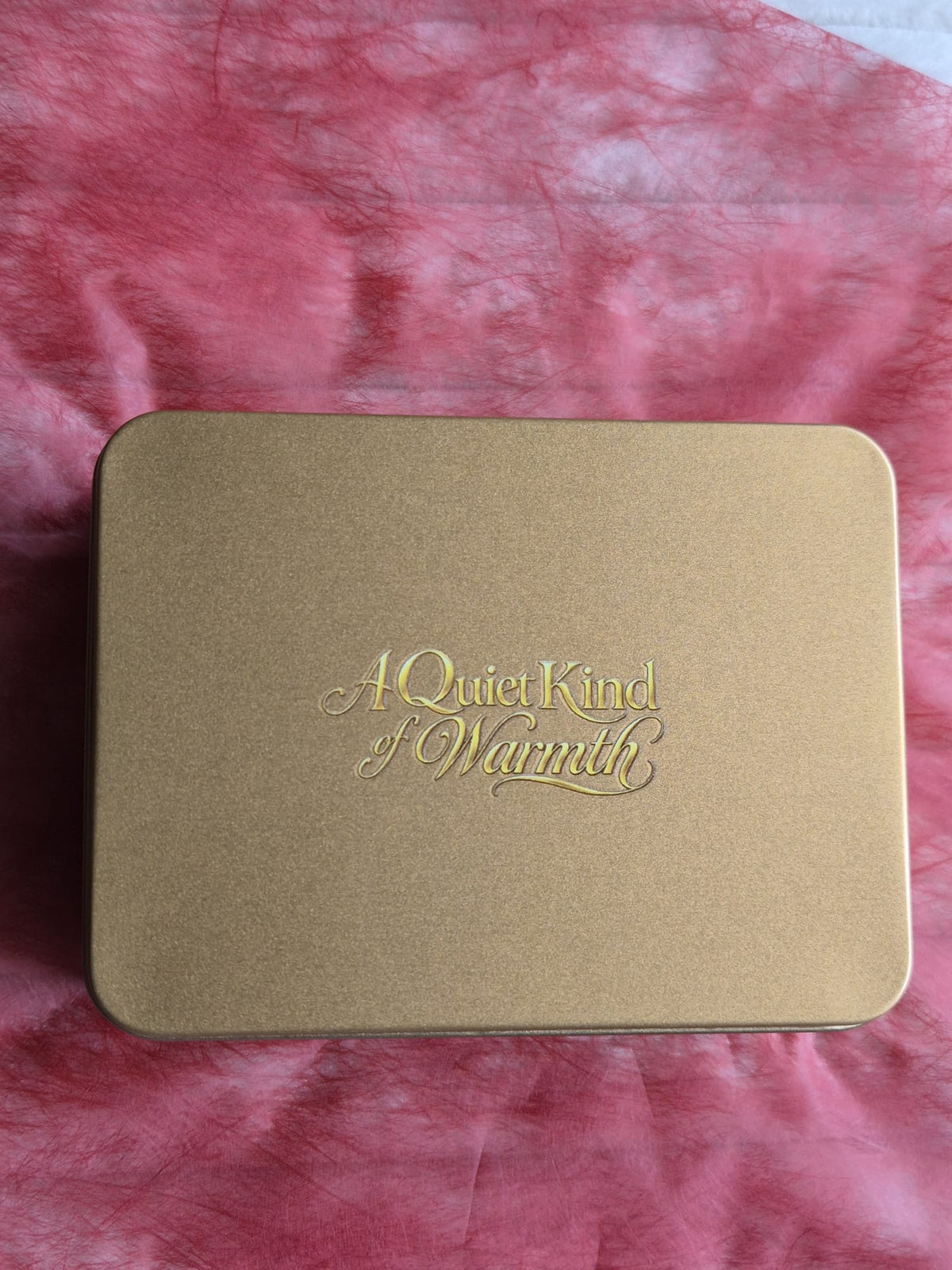 A square metallic gold tin with rounded corners and embossed gold script reading 'A Quiet Kind of Warmth' on the lid, placed on a pink textured fabric background. Thin is full of sweet treats.