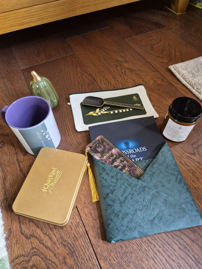 A curated flat-lay of gift items arranged on a dark wood floor, including a teal patterned envelope pouch with a gold tassel, a gold notebook, a purple and white mug, a small green bottle, a black candle jar, and printed cards or a booklet.