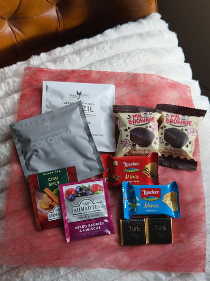 A gift assortment of tea, coffee, chocolates, and snacks arranged on a pink paper sheet over a white blanket, with a brown leather chair visible in the background. 
