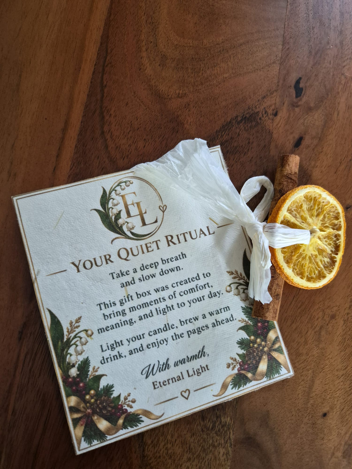 A gift card labeled 'YOUR QUIET RITUAL' with printed text and floral illustrations, wrapped in a white tissue and placed on a wooden surface next to a dried orange slice and a cinnamon stick.