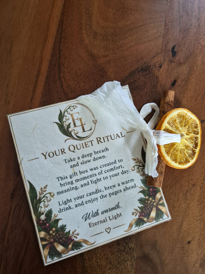 A gift card labeled 'YOUR QUIET RITUAL' with printed text and floral illustrations, wrapped in a white tissue and placed on a wooden surface next to a dried orange slice and a cinnamon stick.