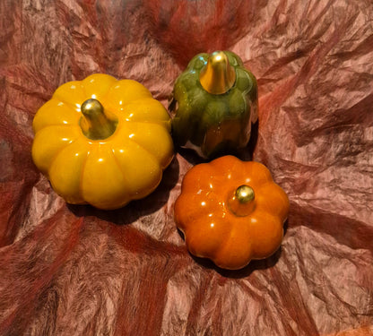 Three small decorative  pumpkins, one yellow, one orange, and one green, each with a metallic gold stem, arranged on a crinkled red-brown paper background.