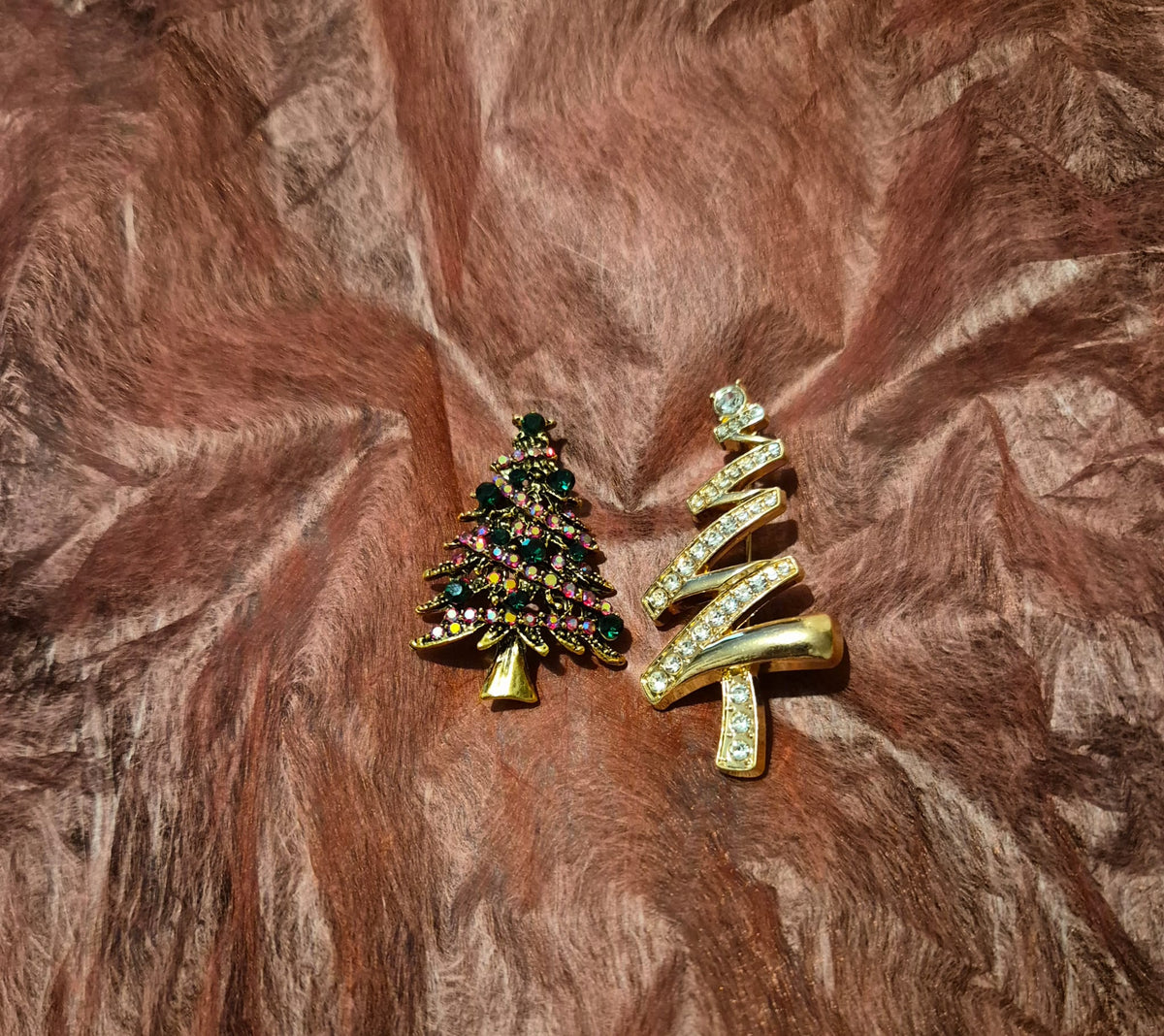 Two Christmas tree-shaped brooches on a brown faux fur background. One brooch is dark green with multicolored rhinestones, and the other is gold with clear rhinestones.
