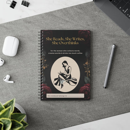 Black and gold spiral notebook for women who love reading and writing, featuring a writer illustration and personalised name space. She Reads, She Writes, She Overthinks journal lying on grey table runner with accesories scattered around.