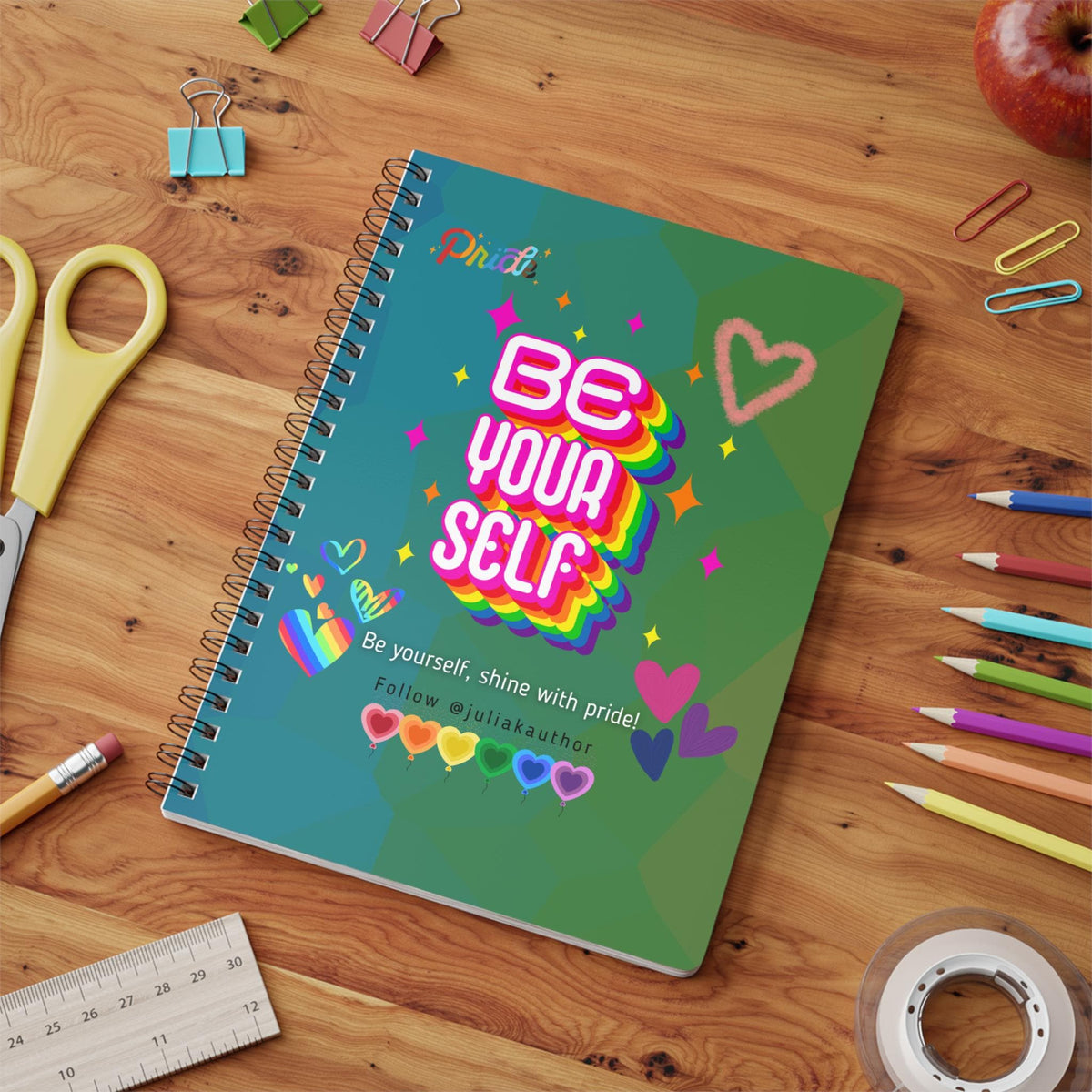 A spiral-bound notebook with a teal and green cover. The cover features the text "BE YOUR SELF" in a rainbow gradient design, along with hearts and stars. The text "Be yourself, shine with pride!" is also visible. Premium Pride month gift.