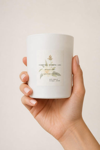 Close-up of a hand holding a luxury white soy wax candle in a matte white glass jar with the Eternal Light label. The candle is scented with gardenia, jasmine and lily and features delicate botanical artwork on a soft neutral background.