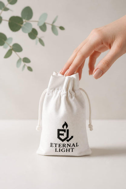 Close-up of a white cotton drawstring gift bag featuring the Eternal Light logo, with a hand placing a luxury candle inside. Styled on a soft neutral background with eucalyptus leaves, the reusable pouch is ideal for elegant gifting and keepsake storage.