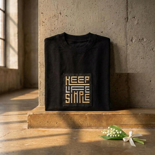 Folded black KEEP LIFE SIMPLE tee on concrete with embroidered gold and silver patch and a little bunch of lily of the valley beside it.
