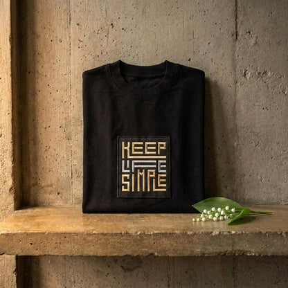 Black heavyweight sweatshirt on its own with bespoke KEEP LIFE SIMPLE embroidered patch in metallic gold and silver, folded on a concrete shelf beside a delicate lily of the valley sprig.
