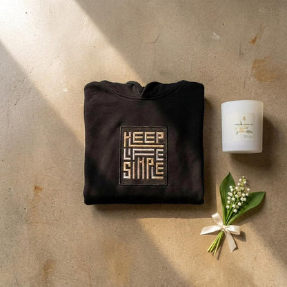 Black heavyweight KEEP LIFE SIMPLE hoodie by Eternal Light on a warm brown concrete shelf, showing the bespoke embroidered patch in metallic gold and silver, styled with one of our candles and a delicate lily of the valley sprig and cream ribbon.