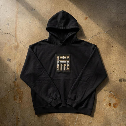 Black heavyweight KEEP LIFE SIMPLE hoodie by Eternal Light lying on a warm brown concrete floor, showing the bespoke embroidered patch in metallic gold and silver, styled with a delicate lily of the valley sprig and cream ribbon.