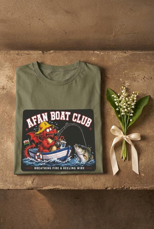 Earth green Afan Boat Club Dragon tee folded on warm brown weathered concrete shelf beside a lily of the valley sprig, red Welsh dragon fishing print centred.