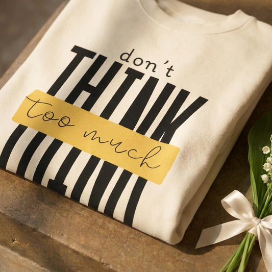 Close-up crop of ecru heavyweight cotton sweatshirt on brown polished concrete shelf, bold black "don't THINK too much" graphic with yellow highlight bar and striped detail filling the frame, lily of the valley sprig with cream ribbon at the edge.