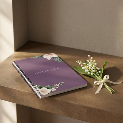 Main product photo of the Eternal Light A5 purple spiral notebook featuring white floral corner illustrations. The word NOTEBOOK is printed in white capital letters in the center, styled on a stone shelf next to a small bouquet of white Lily of the Valley flowers tied with a thin cream ribbon.
