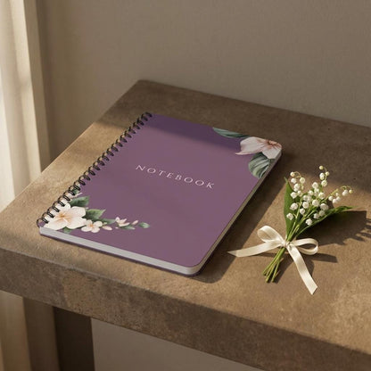 Main product photo of the Eternal Light A5 purple spiral notebook featuring white floral corner illustrations. The word NOTEBOOK is printed in white capital letters in the center, styled on a stone shelf next to a small bouquet of white Lily of the Valley flowers tied with a thin cream ribbon.
