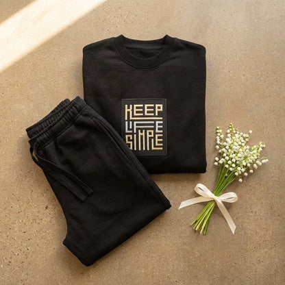 Matching black heavyweight sweatshirt and jogger set with bespoke KEEP LIFE SIMPLE embroidered patch in metallic gold and silver, folded on a concrete floor beside a delicate lily of the valley sprig.