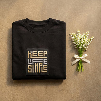 Black heavyweight sweatshirt with bespoke KEEP LIFE SIMPLE embroidered patch in metallic gold and silver, folded on a concrete floor,  beside a delicate lily of the valley sprig.