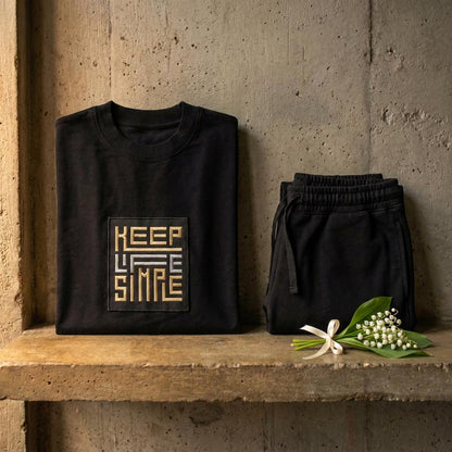 Matching black heavyweight sweatshirt and jogger set with bespoke KEEP LIFE SIMPLE embroidered patch in metallic gold and silver, folded on a concrete shelf beside a delicate lily of the valley sprig.
