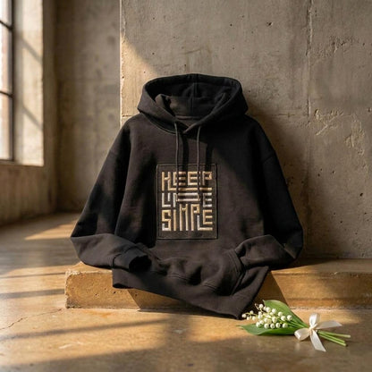 Black heavyweight KEEP LIFE SIMPLE hoodie by Eternal Light on a warm brown concrete shelf, showing the bespoke embroidered patch in metallic gold and silver, styled with a delicate lily of the valley sprig and cream ribbon.