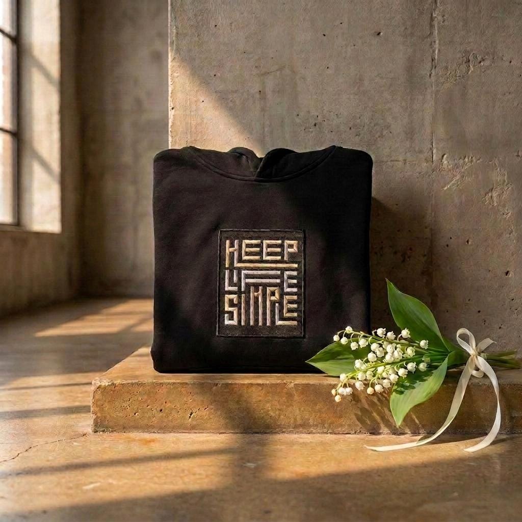 Black heavyweight KEEP LIFE SIMPLE hoodie by Eternal Light on a warm brown concrete shelf, showing the bespoke embroidered patch in metallic gold and silver, styled with a delicate lily of the valley sprig and cream ribbon.