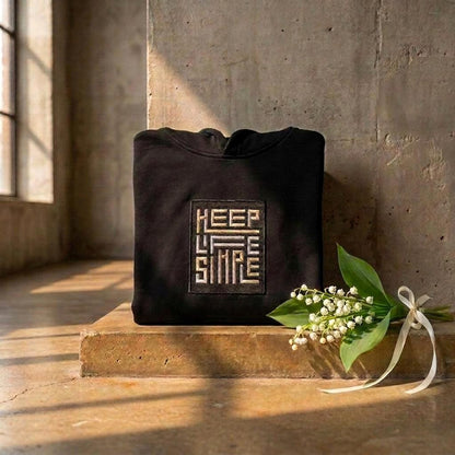 Black heavyweight KEEP LIFE SIMPLE hoodie by Eternal Light on a warm brown concrete shelf, showing the bespoke embroidered patch in metallic gold and silver, styled with a delicate lily of the valley sprig and cream ribbon.