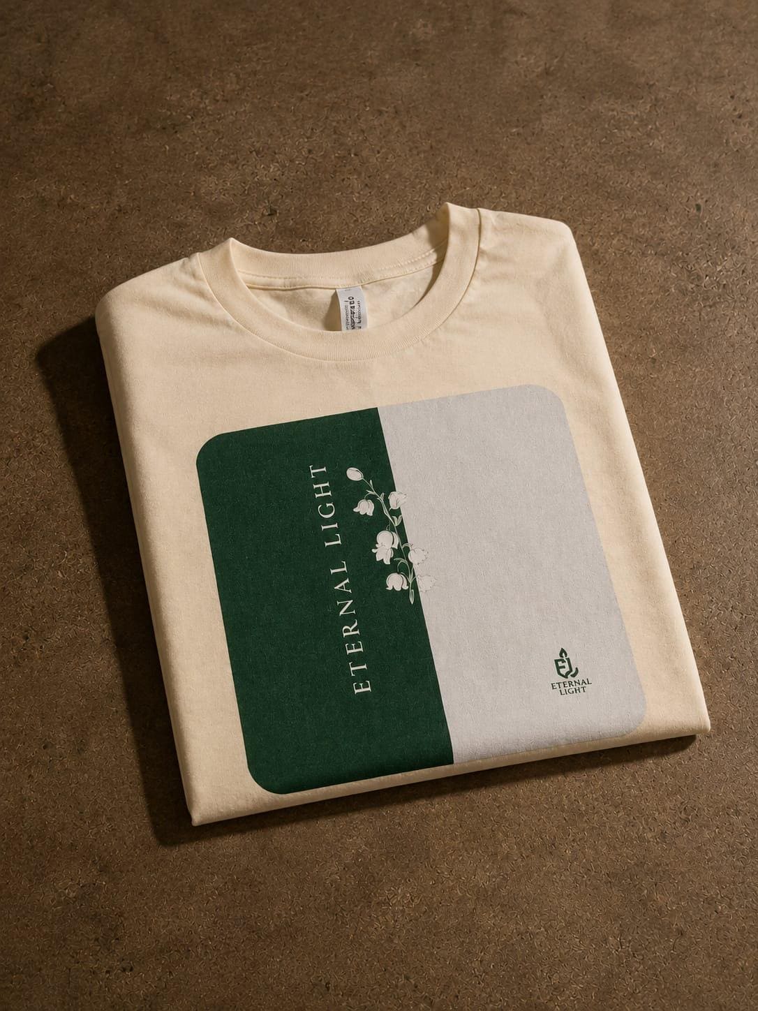 A premium ecru cream t-shirt folded neatly on a textured brown concrete floor, cleanly showcasing the signature Lily of the Valley artwork and Eternal Light logo printed on the front.