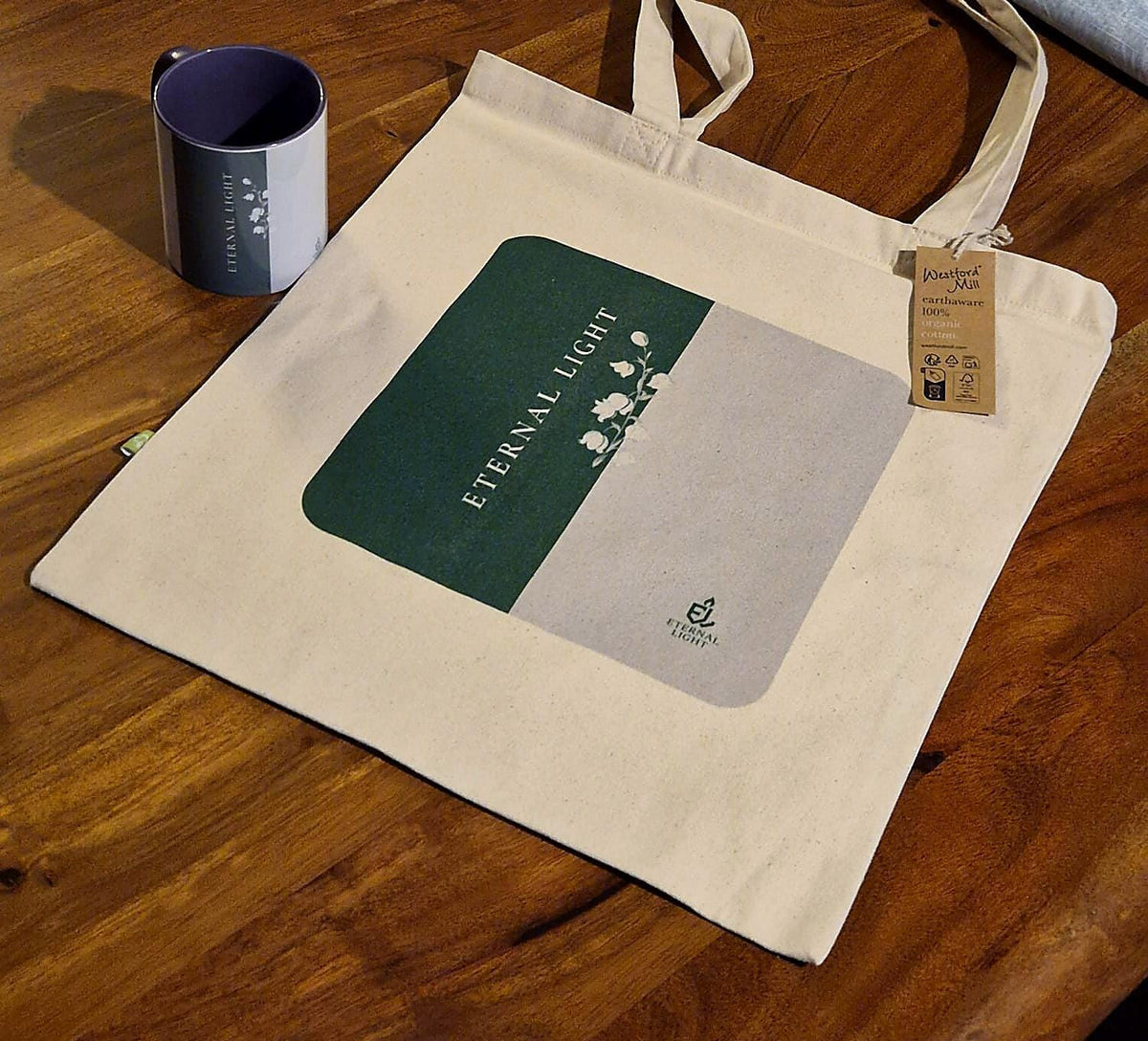 A premium cream EarthAware canvas tote bag laid out on a rustic wooden table next to the matching Eternal Light signature ceramic mug, both featuring the distinctive Lily of the Valley collection artwork.