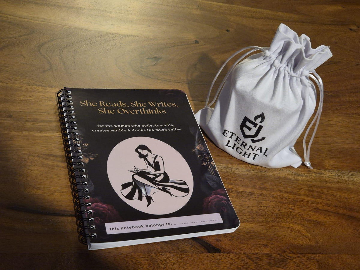 Black and gold spiral notebook for women who love reading and writing, featuring a writer illustration and personalised name space. She Reads, She Writes, She Overthinks journal. Notebook lying on a wooden table with our luxury candle inside its cotton bag beside it.