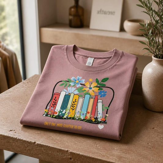 Aesthetic studio photo of the Hazy Pink graphic t-shirt neatly folded on a textured stone table. The print shows a colourful floral book stack inside an audiobook music interface with the text Only One More Chapter To Go, styled next to a rustic ceramic vase.