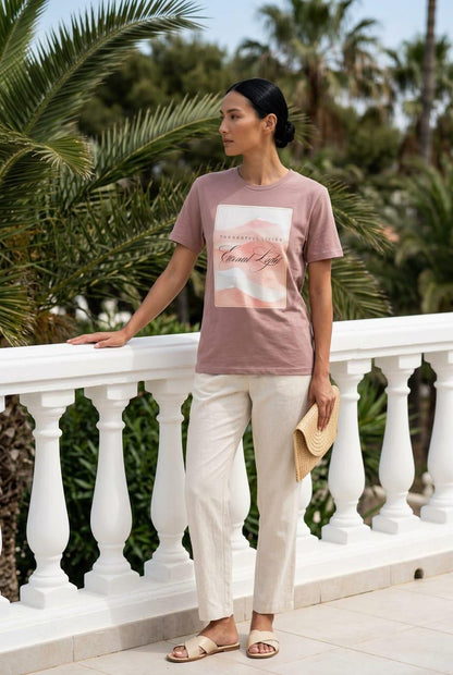 Elegant lookbook lifestyle photo of a model standing by a white balustrade terrace with palm trees, wearing the Eternal Light Hazy Pink graphic t-shirt. The tee showcases the minimalist pastel watercolor Thoughtful Living graphic, styled beautifully with cream linen trousers, neutral leather cross-strap sandals, and a woven straw clutch bag.