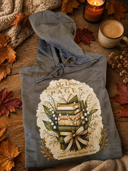 An extra-heavyweight unisex hoodie in a rich olive green colour, shown in an autumnal background. The chest features an elegant, hand-drawn dark academia graphic showcasing a stack of vintage books intertwined with delicate white Lily of the Valley blossoms. Ornate script reads, "She Lives Between Pages Where Stories Become Home."
