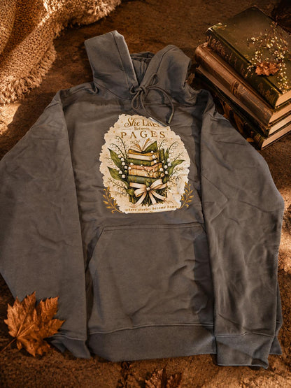 An extra-heavyweight unisex hoodie in a rich olive green colour, shown lying flat in an autumnal background next to a stack of books. The chest features an elegant, hand-drawn dark academia graphic showcasing a stack of vintage books intertwined with delicate white Lily of the Valley blossoms. Ornate script reads, "She Lives Between Pages Where Stories Become Home."