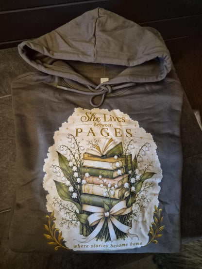 An extra-heavyweight unisex hoodie in a rich olive green colour, nicely folded on top of an ottoman. The chest features an elegant, hand-drawn dark academia graphic showcasing a stack of vintage books intertwined with delicate white Lily of the Valley blossoms. Ornate script reads, "She Lives Between Pages Where Stories Become Home."