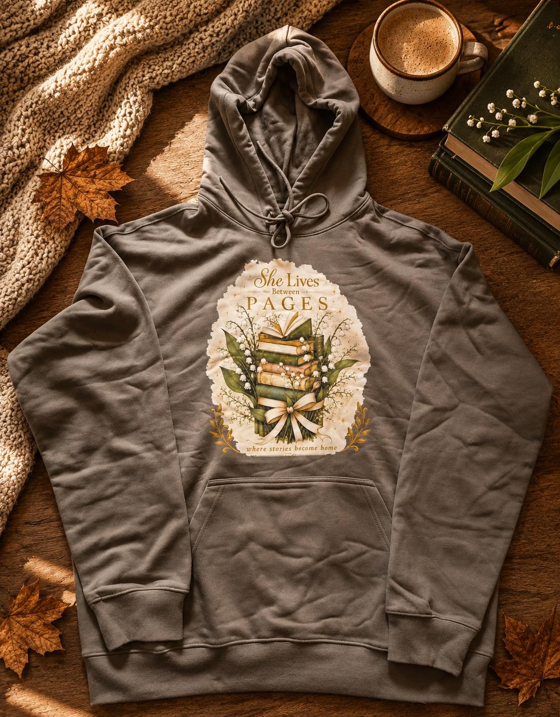 An extra-heavyweight unisex hoodie in a rich olive green colour, shown lying flat in an autumnal background next to a stack of books. The chest features an elegant, hand-drawn dark academia graphic showcasing a stack of vintage books intertwined with delicate white Lily of the Valley blossoms. Ornate script reads, "She Lives Between Pages Where Stories Become Home."