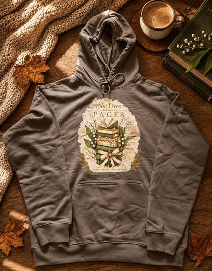 An extra-heavyweight unisex hoodie in a rich olive green colour, shown lying flat in an autumnal background next to a stack of books. The chest features an elegant, hand-drawn dark academia graphic showcasing a stack of vintage books intertwined with delicate white Lily of the Valley blossoms. Ornate script reads, "She Lives Between Pages Where Stories Become Home."