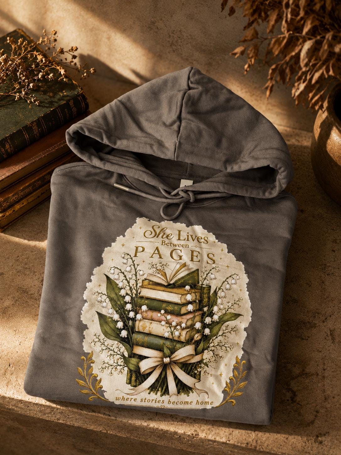 An extra-heavyweight unisex hoodie in a rich olive green colour, shown on a concrete shelf in an autumnal background. The chest features an elegant, hand-drawn dark academia graphic showcasing a stack of vintage books intertwined with delicate white Lily of the Valley blossoms. Ornate script reads, "She Lives Between Pages Where Stories Become Home."