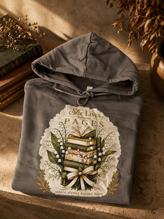 An extra-heavyweight unisex hoodie in a rich olive green colour, shown on a concrete shelf in an autumnal background. The chest features an elegant, hand-drawn dark academia graphic showcasing a stack of vintage books intertwined with delicate white Lily of the Valley blossoms. Ornate script reads, "She Lives Between Pages Where Stories Become Home."