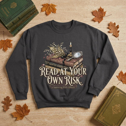A premium 370GSM heavyweight unisex sweatshirt in a unique charcoal espresso brown, laid flat on a beige stone surface. The front displays a detailed dark academia design featuring a stack of vintage books titled "Gothic Tales." A burning black candle, delicate lily of the valley blossoms, and a vintage cracked mirror rest atop the books. Cream text boldly reads "READ AT YOUR OWN RISK." The cozy arrangement is framed by rustic, dried autumn oak leaves and antique library volumes.