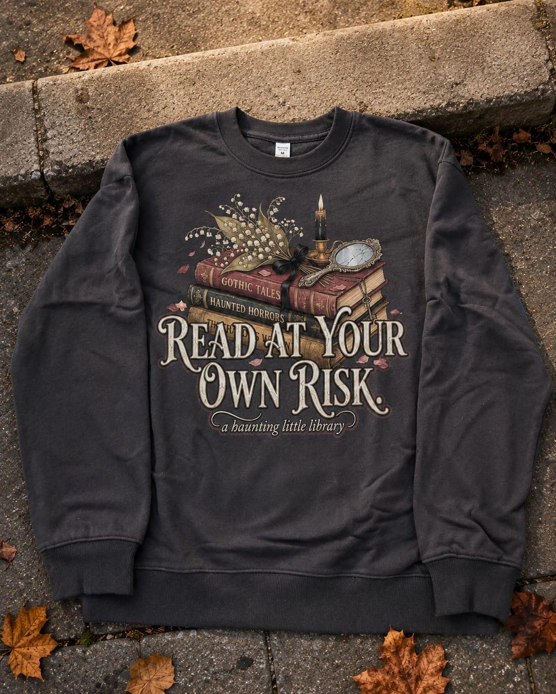 A premium 370GSM heavyweight unisex sweatshirt in a unique charcoal espresso brown, laid flat on a beige stone kerb. The front displays a detailed dark academia design featuring a stack of vintage books titled "Gothic Tales." A burning black candle, delicate lily of the valley blossoms, and a vintage cracked mirror rest atop the books. Cream text boldly reads "READ AT YOUR OWN RISK." The cozy arrangement is framed by rustic, dried autumn oak leaves.