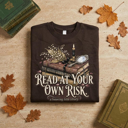 A premium 370GSM heavyweight unisex sweatshirt in a unique charcoal espresso brown, neatly folded on a beige stone surface. The front displays a detailed dark academia design featuring a stack of vintage books titled "Gothic Tales." A burning black candle, delicate lily of the valley blossoms, and a vintage cracked mirror rest atop the books. Cream text boldly reads "READ AT YOUR OWN RISK." The cozy arrangement is framed by rustic, dried autumn oak leaves and antique library volumes.