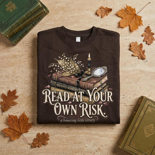 A premium 370GSM heavyweight unisex sweatshirt in a unique charcoal espresso brown, neatly folded on a beige stone surface. The front displays a detailed dark academia design featuring a stack of vintage books titled "Gothic Tales." A burning black candle, delicate lily of the valley blossoms, and a vintage cracked mirror rest atop the books. Cream text boldly reads "READ AT YOUR OWN RISK." The cozy arrangement is framed by rustic, dried autumn oak leaves and antique library volumes.