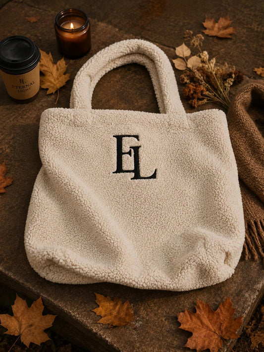 Natural cream borg tote bag with black embroidered EL monogram styled on a warm brown surface with autumn leaves and soft natural light.