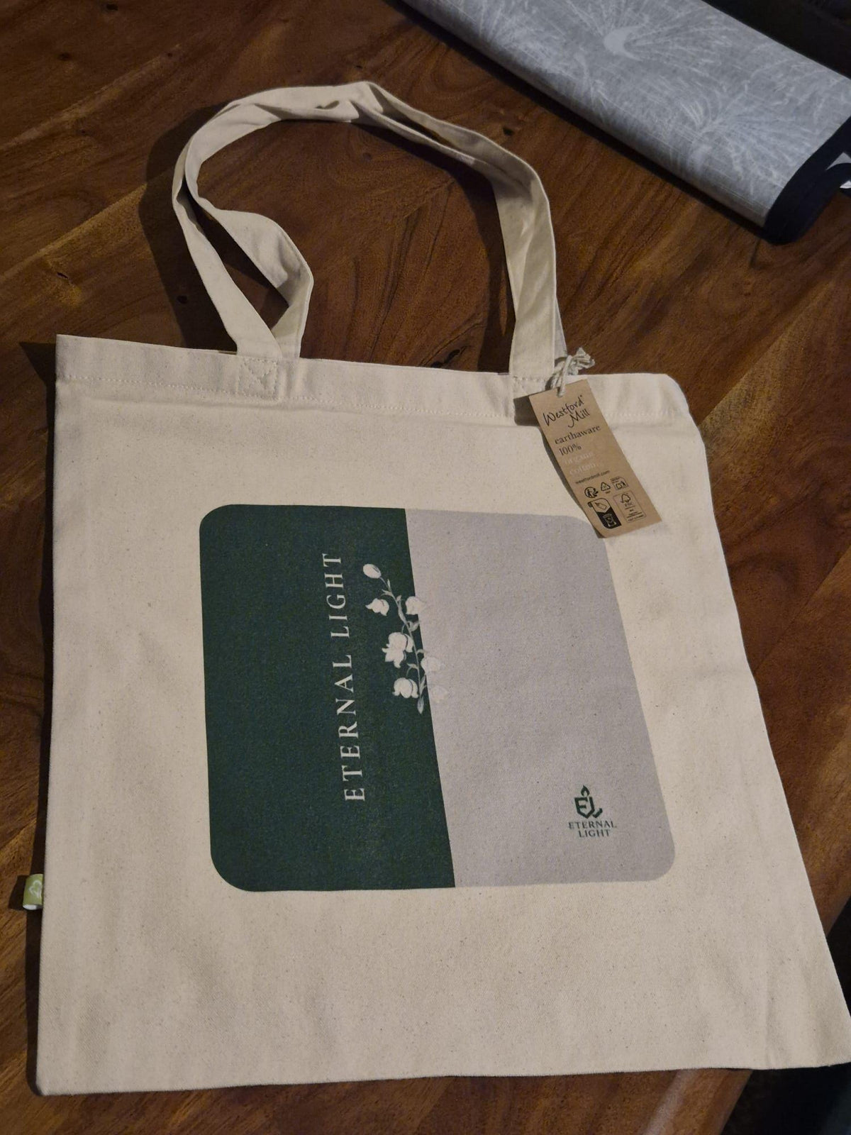 A premium cream EarthAware canvas tote bag laid out on a rustic wooden table featuring the distinctive Lily of the Valley collection artwork.