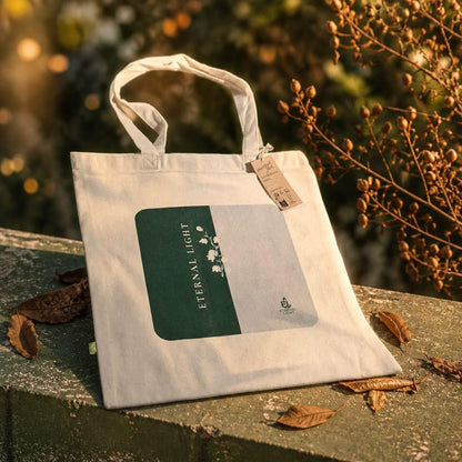 A premium cream EarthAware canvas organic tote bag for life displayed on a stone ledge in warm golden sunlight, showcasing the signature split green and cream print with the Eternal Light wordmark and Lily of the Valley illustration, surrounded by dry leaves.