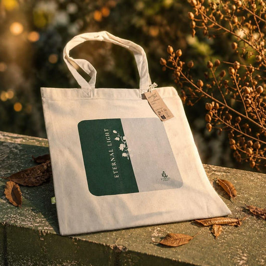 A premium cream EarthAware canvas organic tote bag for life displayed on a stone ledge in warm golden sunlight, showcasing the signature split green and cream print with the Eternal Light wordmark and Lily of the Valley illustration, surrounded by dry leaves.