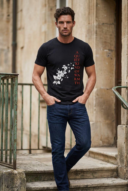 Outdoor lifestyle lookbook photo of a man walking down ancient stone building steps, wearing the Eternal Light black unisex graphic t-shirt. The tee features fine white botanical line work and vertical crimson red typography reading A Quiet Kind of Warmth, styled elegantly with classic dark blue denim jeans.